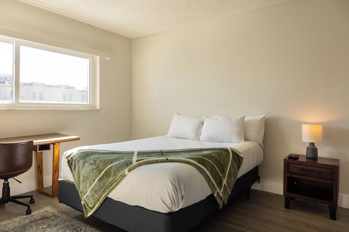A bedroom with a bed, a desk, a chair, a nightstand, and a window.