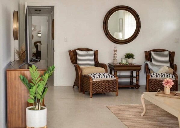 A living room with two wicker chairs, a round mirror, a small table with a lamp, and a potted plant.