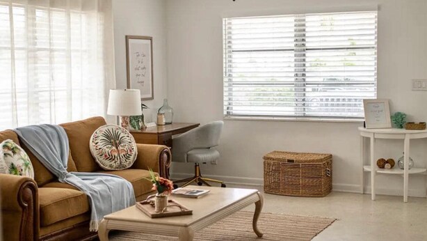 Un salon avec un canapé marron, une table basse blanche, un fauteuil gris, un panier en osier et une table d’appoint blanche.