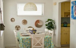 Uma área de refeições com uma mesa posta para quatro pessoas, rodeada por cadeiras brancas, uma luminária pendente trançada e decorações nas paredes.