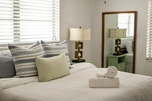Cama bem arrumada com almofadas listradas e lisas, luminária de cabeceira e espelho sobre um criado-mudo verde.