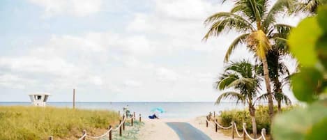 A beach path with a lifeguard tower, palm trees, and a person under an umbrella.