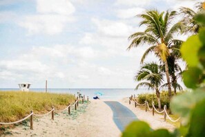 Een strandpad met een redershut, palmbomen en iemand onder een parasol.