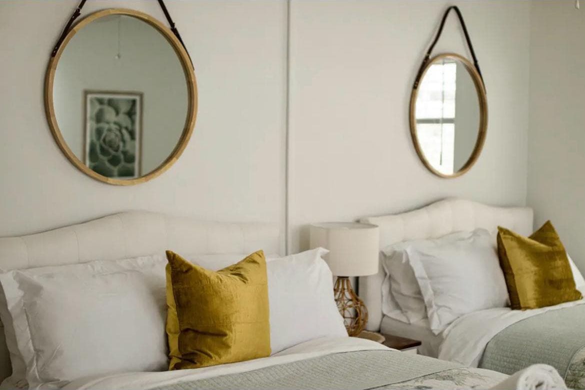 A hotel room with two beds, each with white bedding and gold pillows. There are two round mirrors on the walls, and a framed picture is visible on the left mirror.