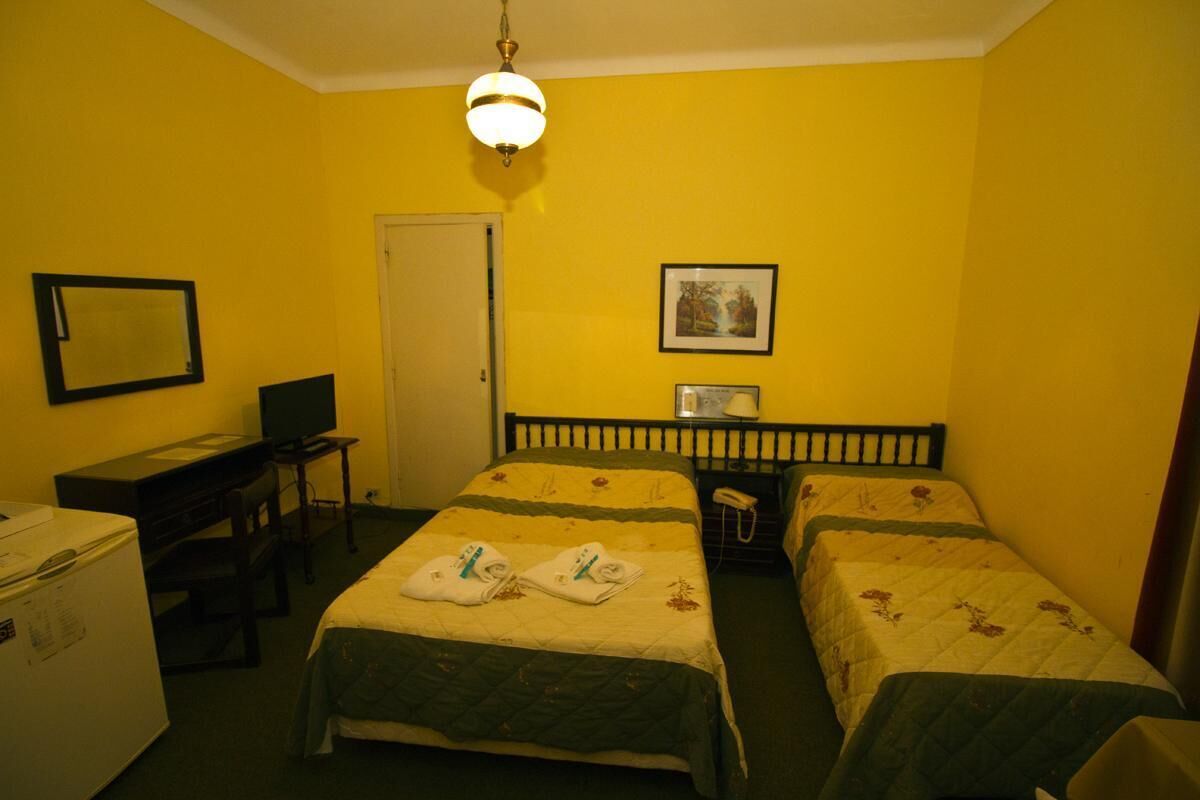 A hotel room with two single beds, a desk with a computer, a refrigerator, and a hanging light fixture.