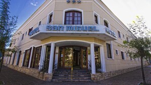 Un hotel llamado "San Miguel" con un estilo arquitectónico clásico, que presenta una entrada destacada con escalones y columnas, y un cielo despejado al fondo.