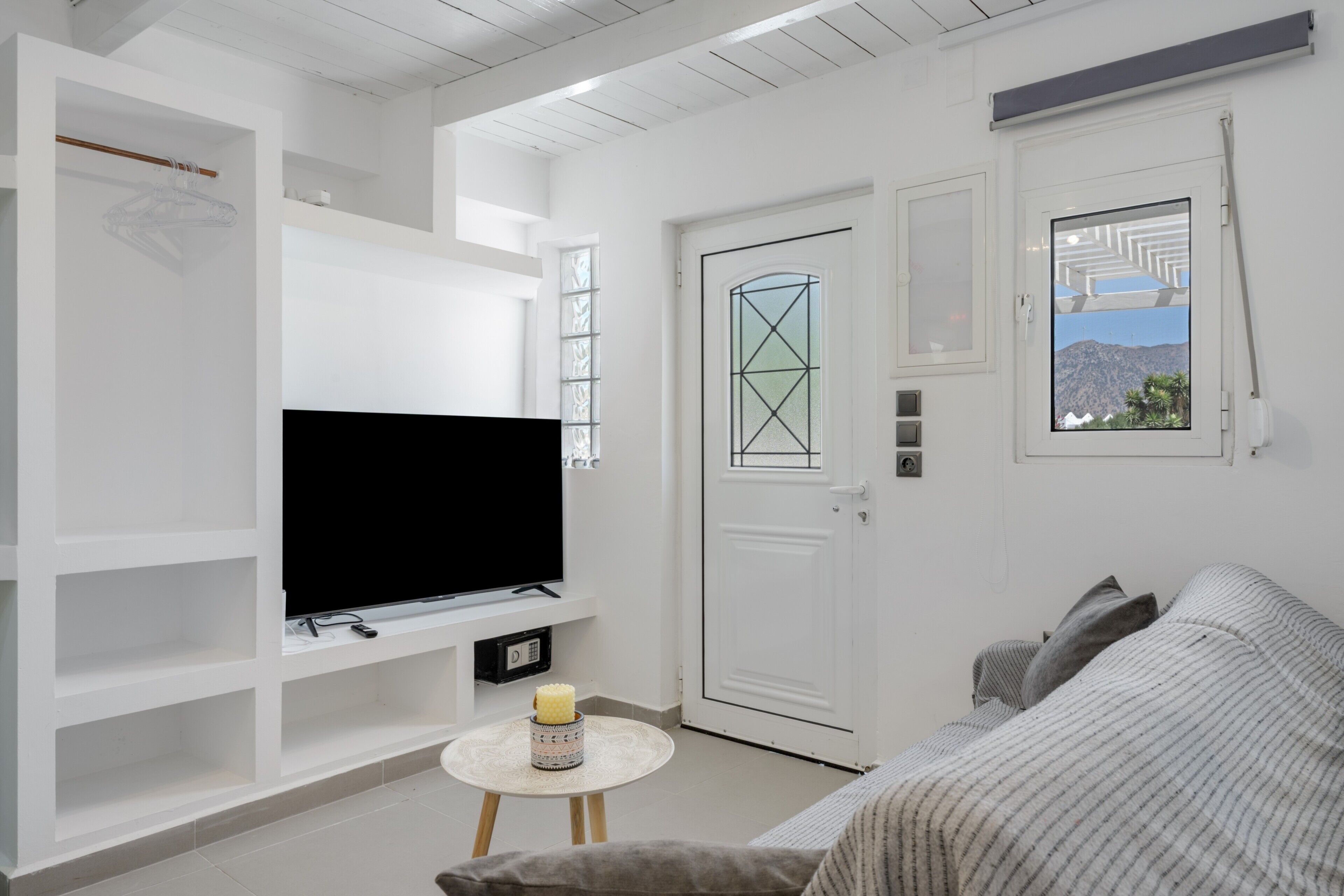 A room with a television on a white shelf, a door with a window, a small round table with a candle, and a sofa.