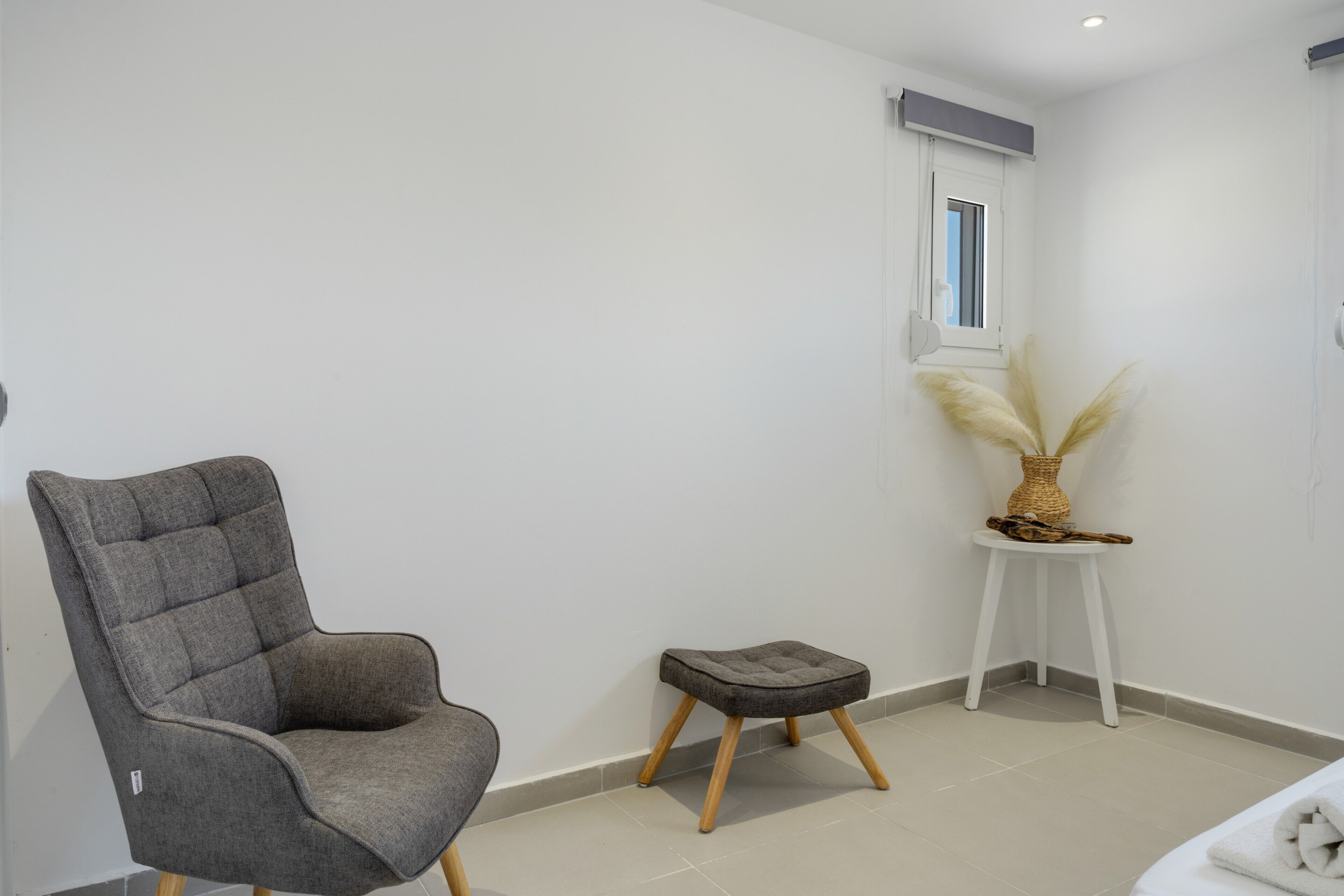 A modern room with a grey upholstered armchair, a small stool, and a decorative vase on a white side table.