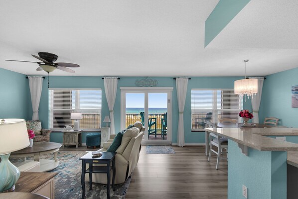 A spacious living room with a view of the ocean, a ceiling fan, and a dining area with a kitchen counter.