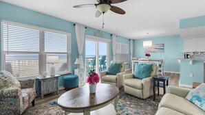 A living room with a ceiling fan, two windows with blinds, a round coffee table with a vase, and a view of the ocean through the windows. - 🌊Gulf View-Steps to Beach-Heated Pool-Islander Beach Resort 2002🌅 (Fort Walton Beach)