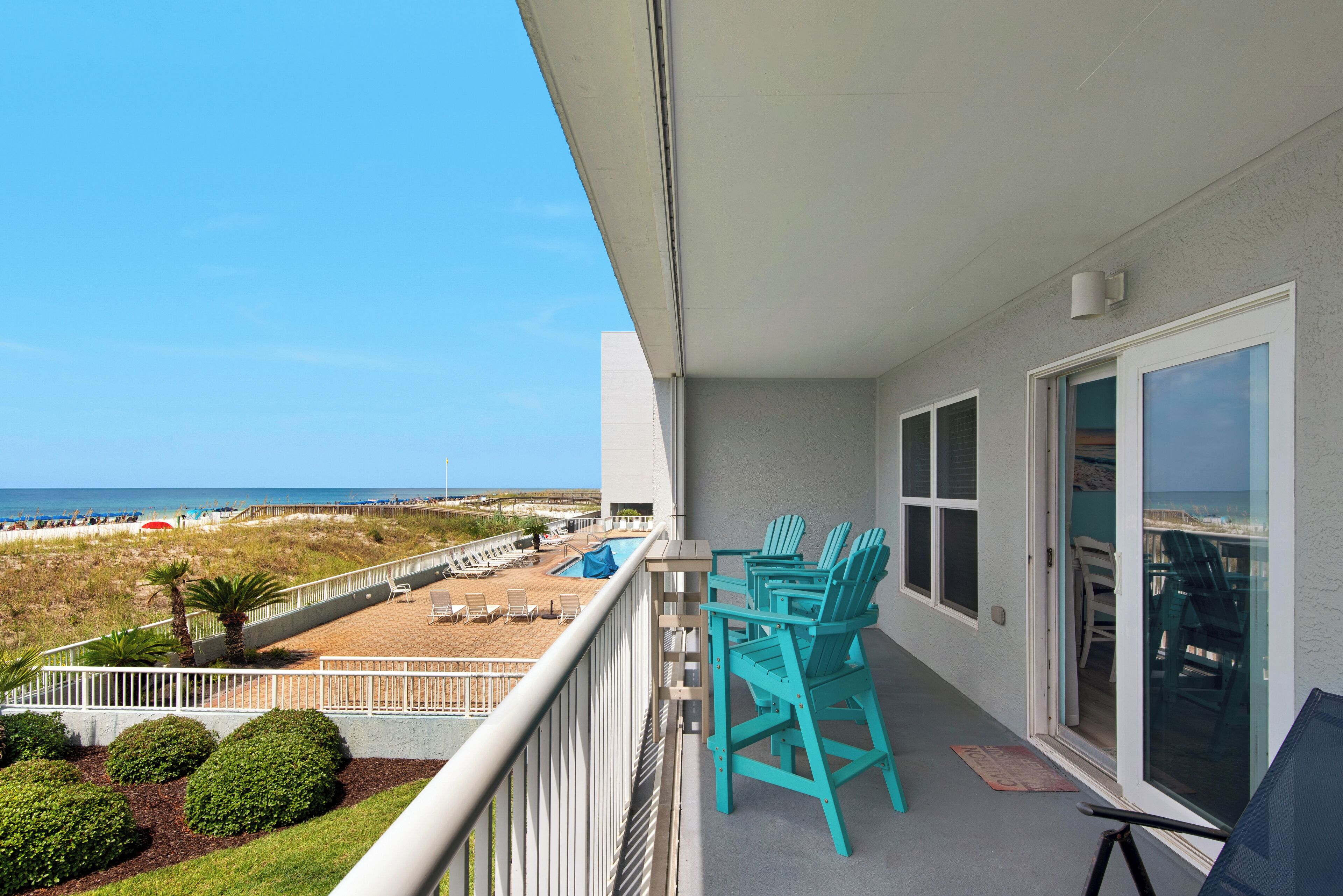 A balcony with turquoise chairs, a view of the beach, and a pool area.