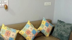 Un canapé beige agrémenté de coussins aux motifs colorés, et un mur uni sur lequel est accrochée une seule œuvre d’art encadrée, au-dessus d’une prise de courant.
