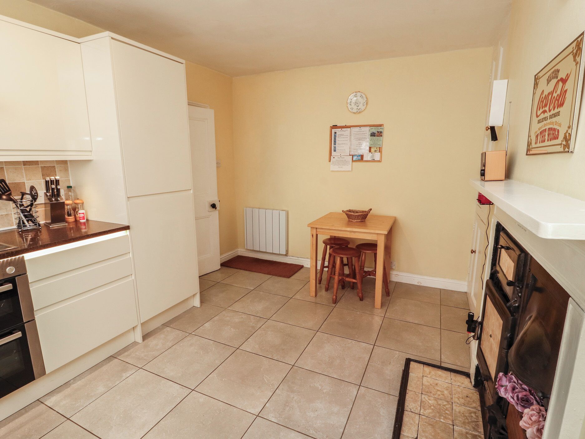 A compact kitchen with white cabinets, a wooden table with stools, a radiator, and a wall-mounted clock.