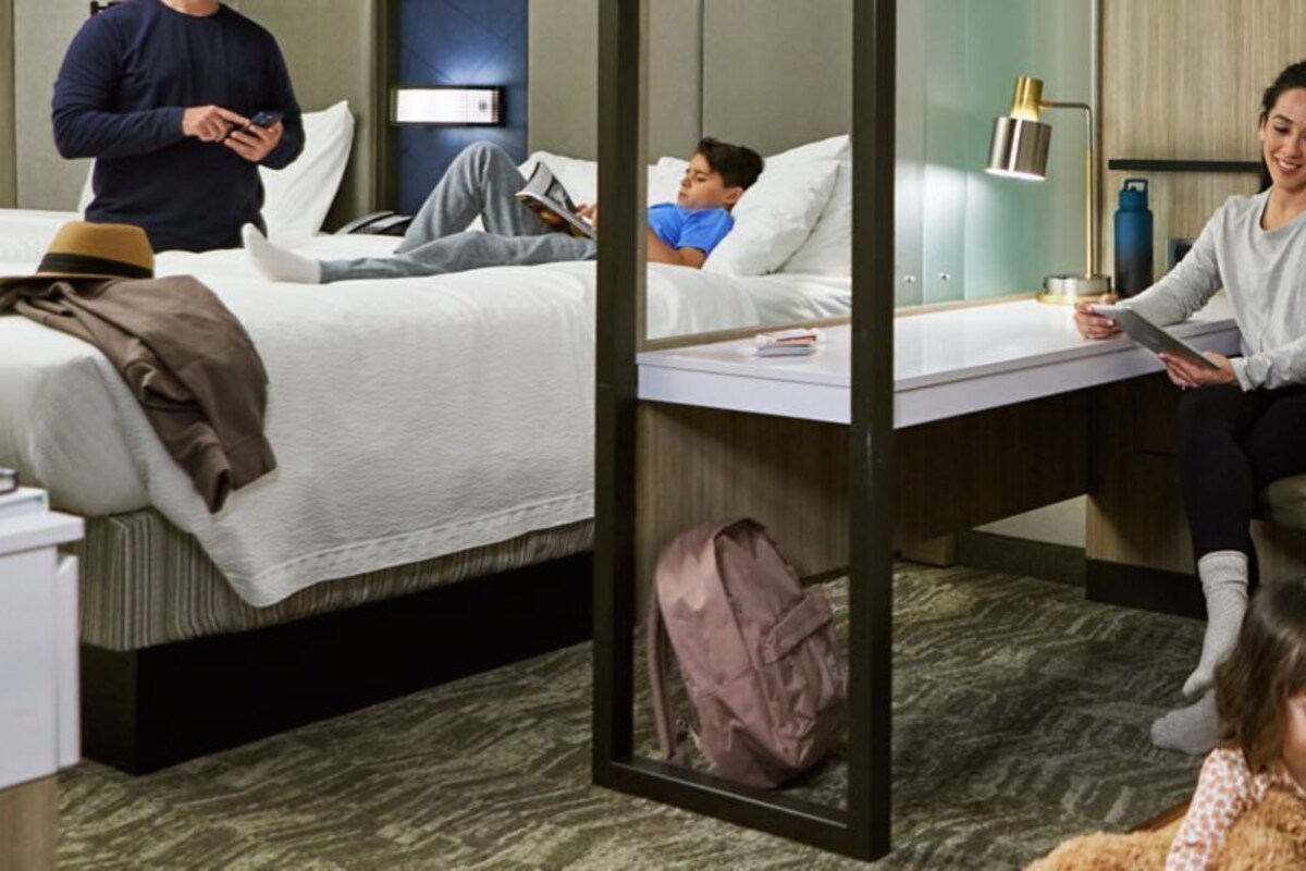 Une chambre d’hôtel avec un lit, un bureau et un enfant qui joue à des jeux vidéo.