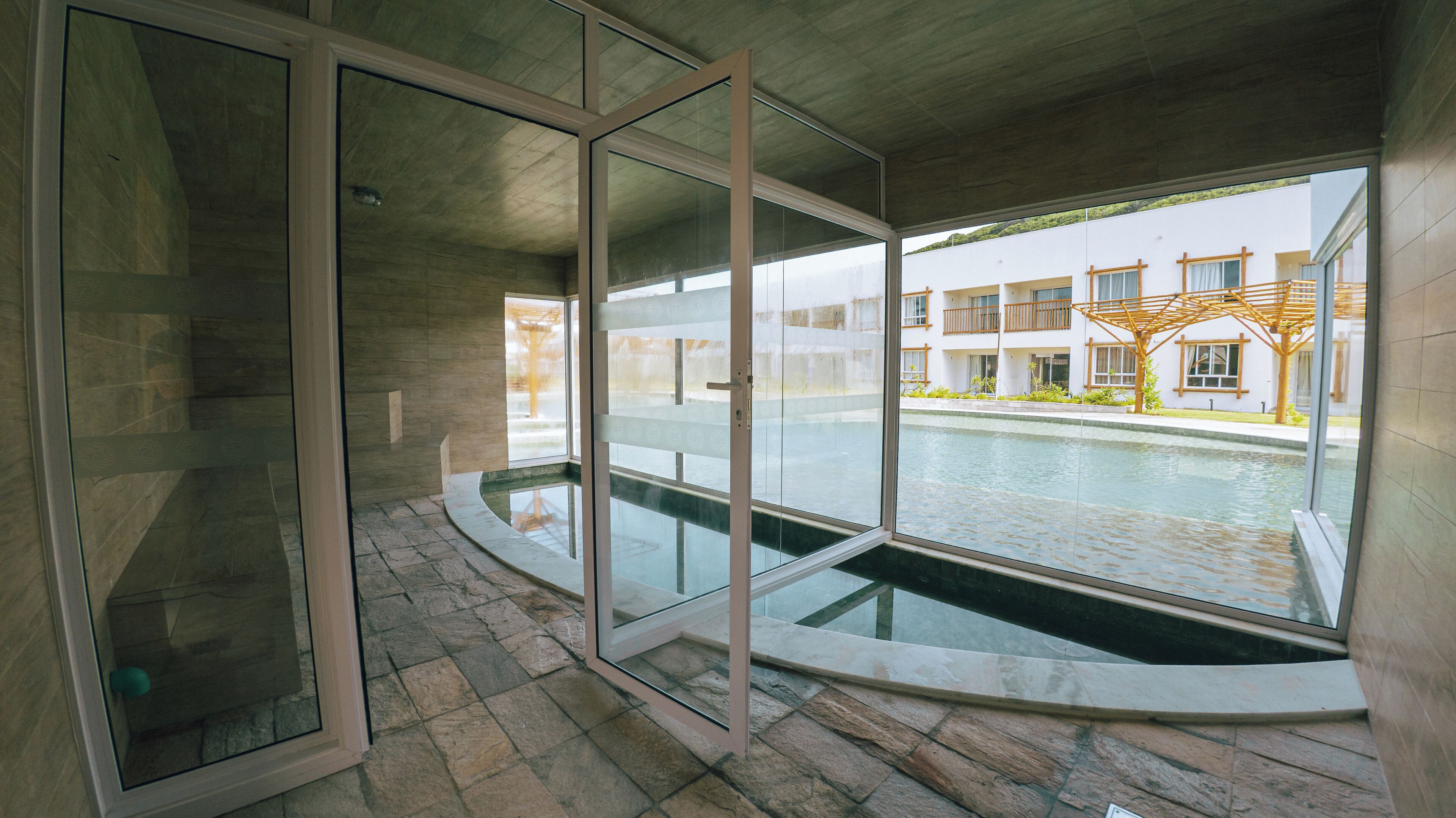 Ein modernes Interieur mit einem von Glas umgebenen Pool, Steinböden und Blick auf ein Wohngebäude.