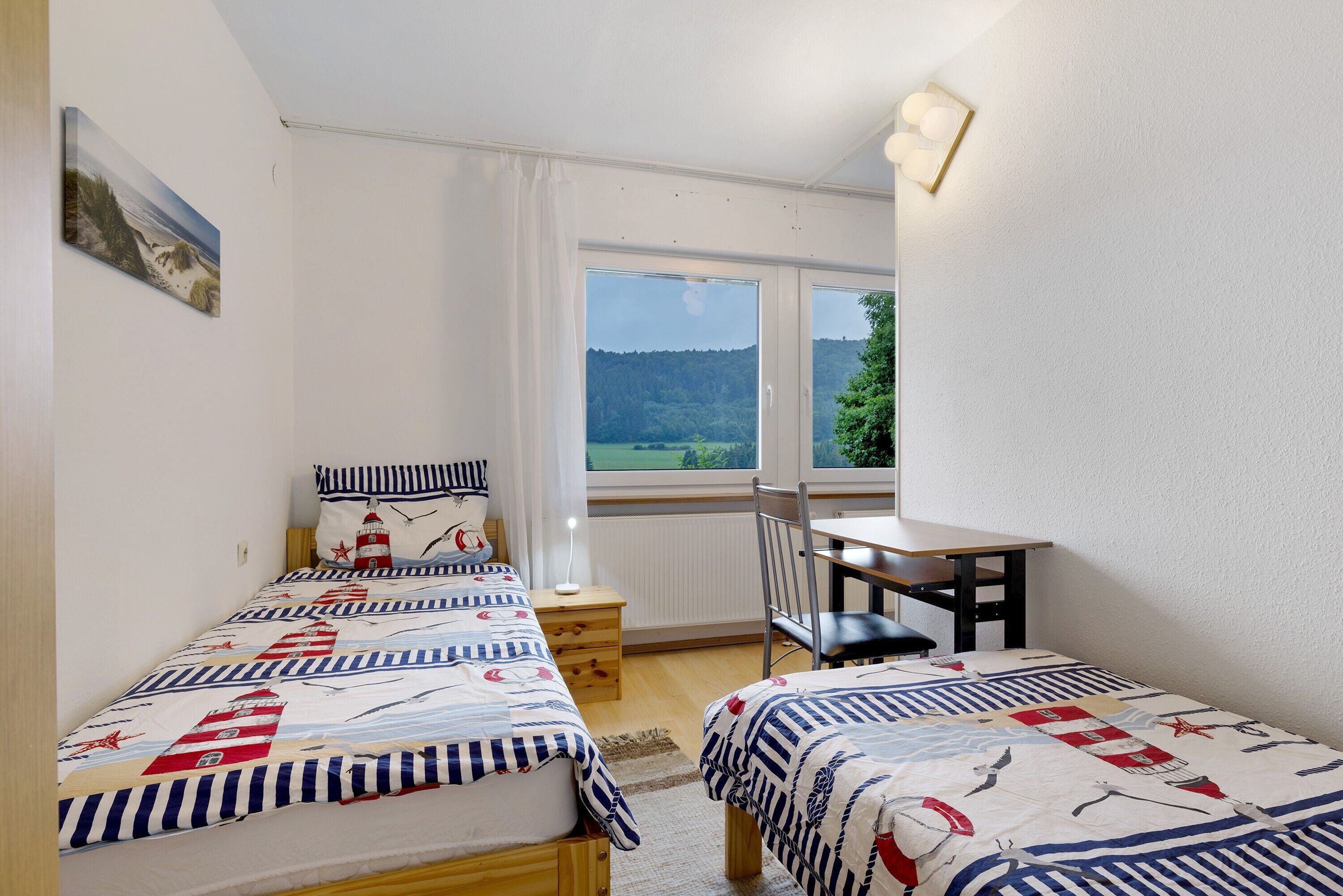 A bedroom with two beds, a desk, and a view of a forested landscape.