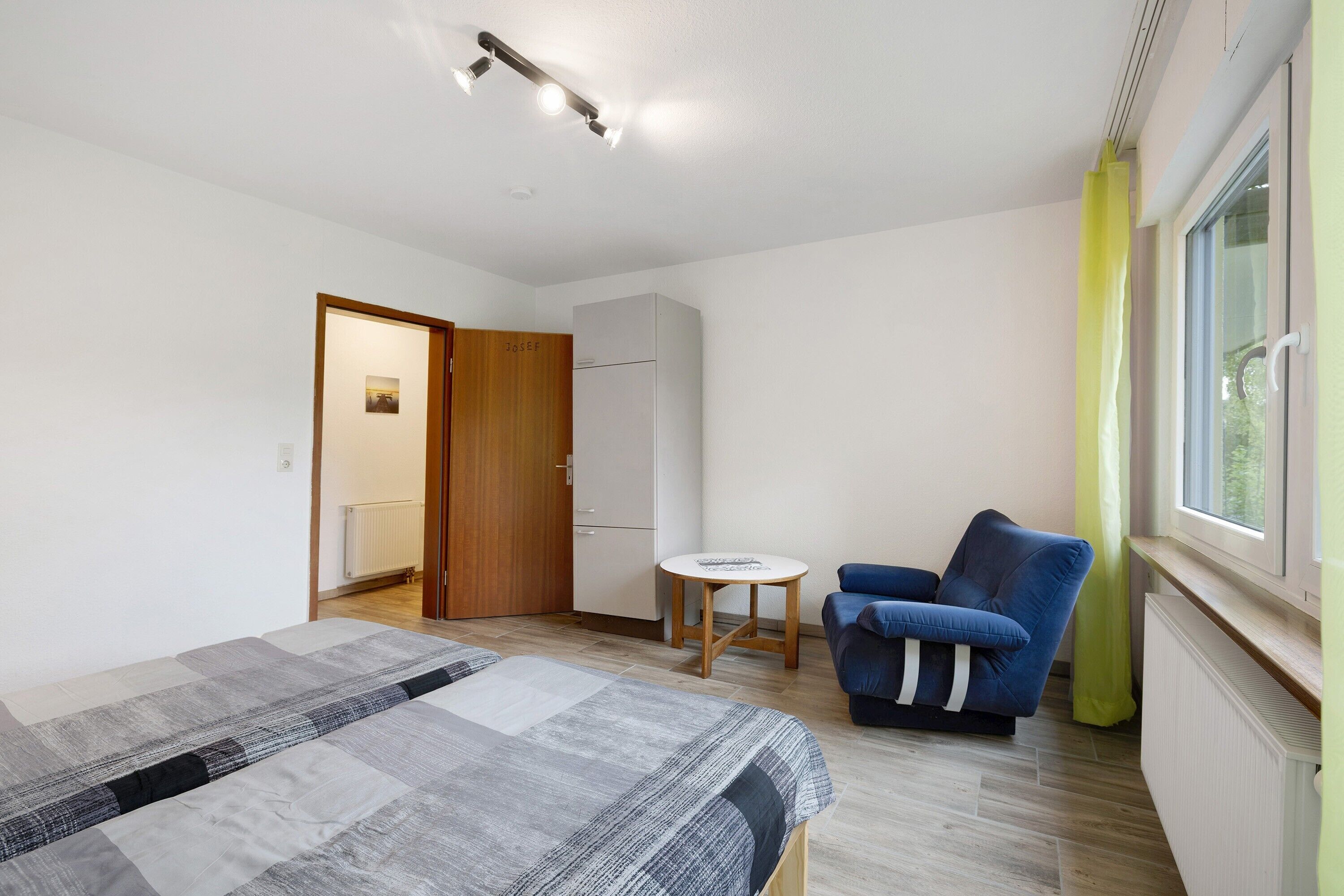 A bedroom with a bed, a blue chair, a small table, a wardrobe, and a window with curtains.
