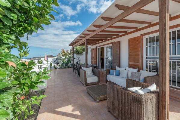 Une terrasse couverte, aménagée avec des meubles en osier, un canapé blanc et un coin repas comprenant des chaises et une table.