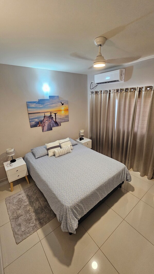Une chambre à coucher bien rangée, avec un lit, des tables de chevet, un climatiseur fixé au mur et un tableau représentant un coucher de soleil.