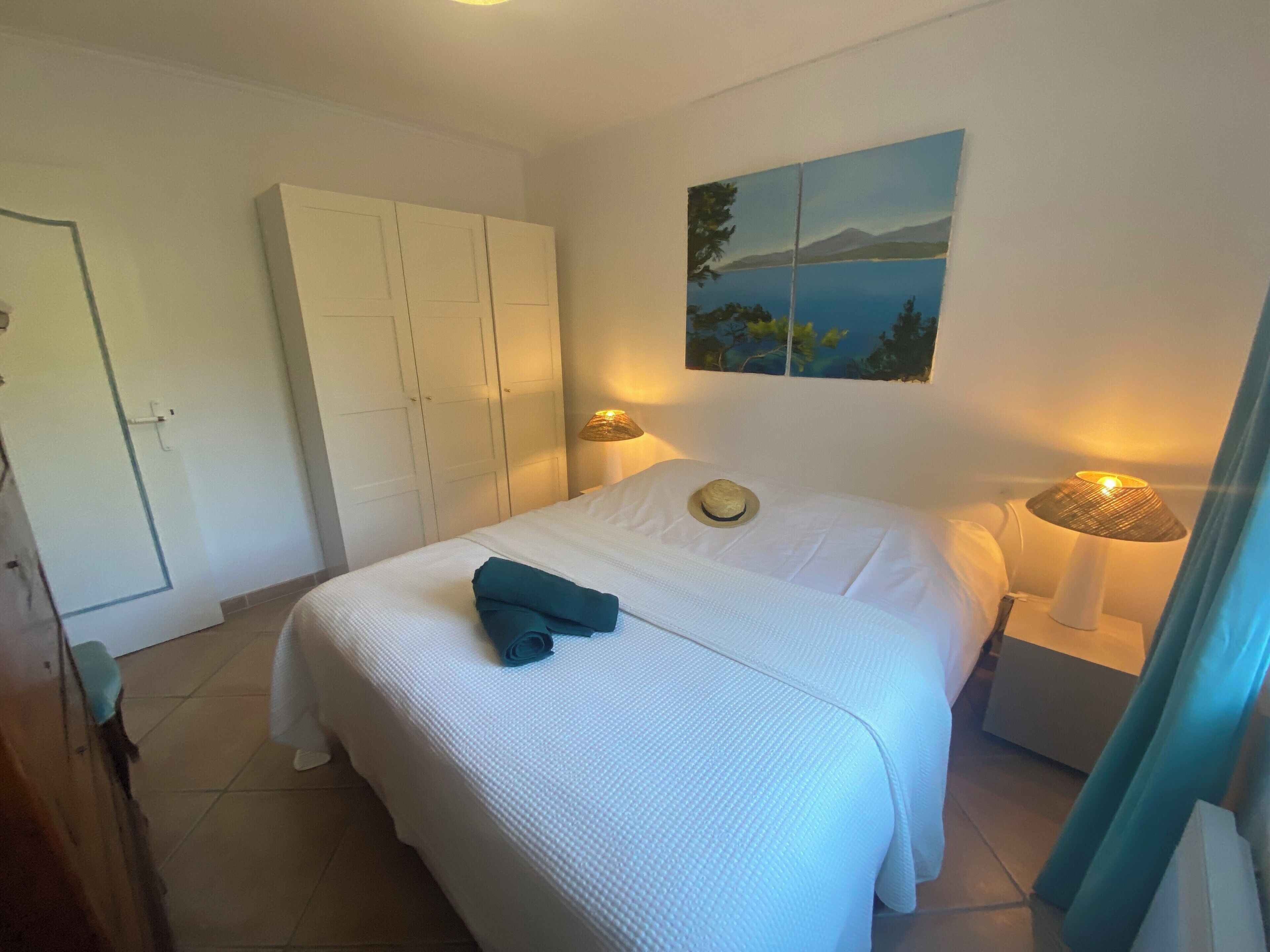 Un letto rifatto con cura, un asciugamano arrotolato, un quadro che raffigura un paesaggio e una lampada su un comodino.