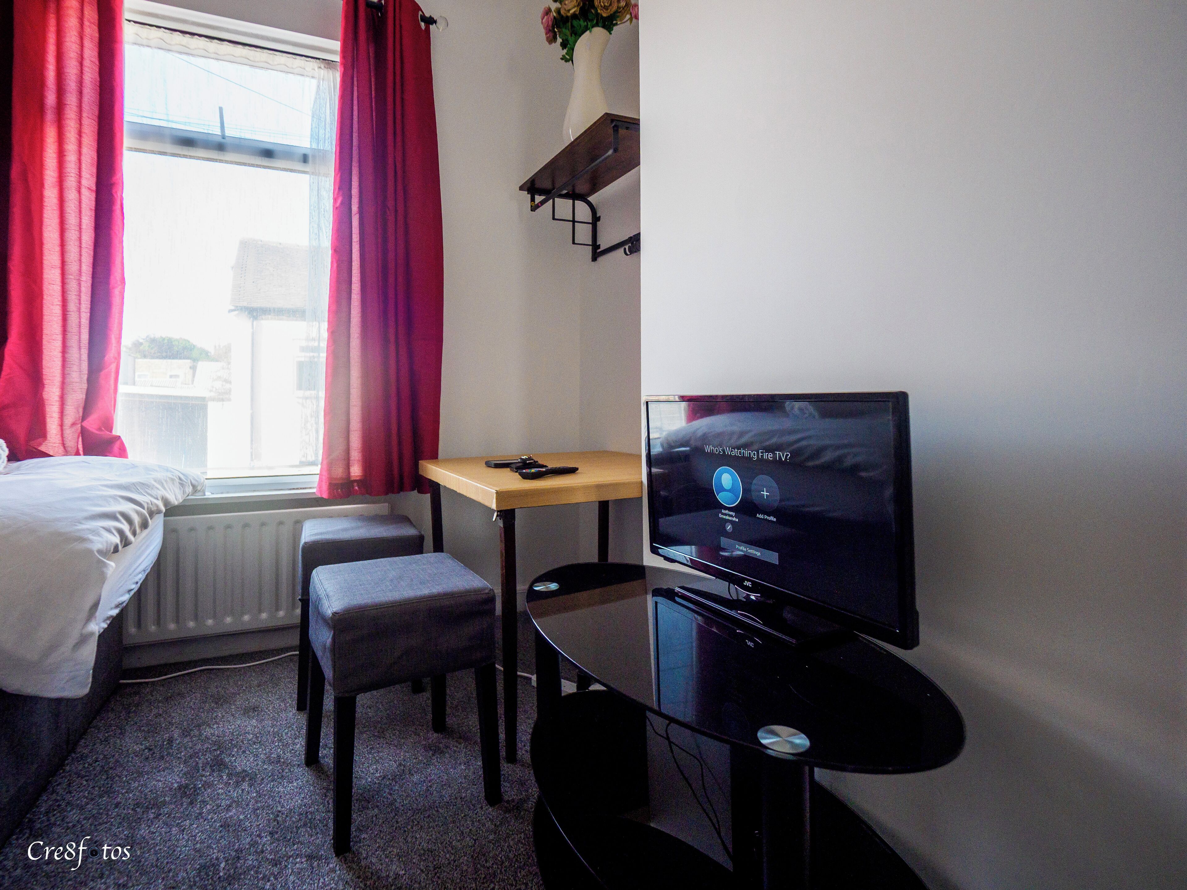 A small hotel room with a bed, a desk, a TV, and a window with red curtains.