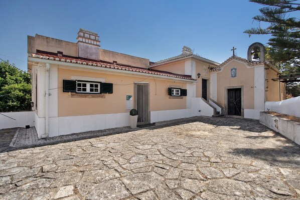 A building with a stone-paved courtyard, a small bell tower, and a door with a cross above it.