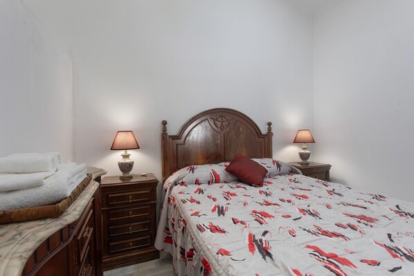 A bedroom with a wooden headboard, two bedside tables with lamps, a bed with a patterned bedspread, and a dresser with folded towels.