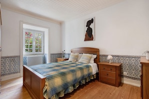 Une chambre à coucher avec un lit en bois, une table de chevet, une commode, une fenêtre donnant sur des fleurs et un tableau accroché au mur.