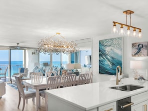 A modern kitchen with a central island, a dining area with chairs, a chandelier, and artwork on the walls.
