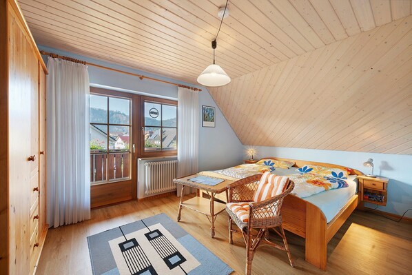 A bedroom with a sloped ceiling, a bed with a floral bedspread, a wicker chair, a table, and a view of the outdoors through a window. - Apartment 'Panoramablick' with Mountain View, Shared Terrace and Wi-Fi (Haslach im Kinzigtal)