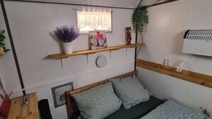 A compact room with a bed, a small window with a curtain, a wall-mounted air conditioner, and a shelf with decorative items. - Construction trailer on the farm in the Weserbergland (Coppenbrügge)