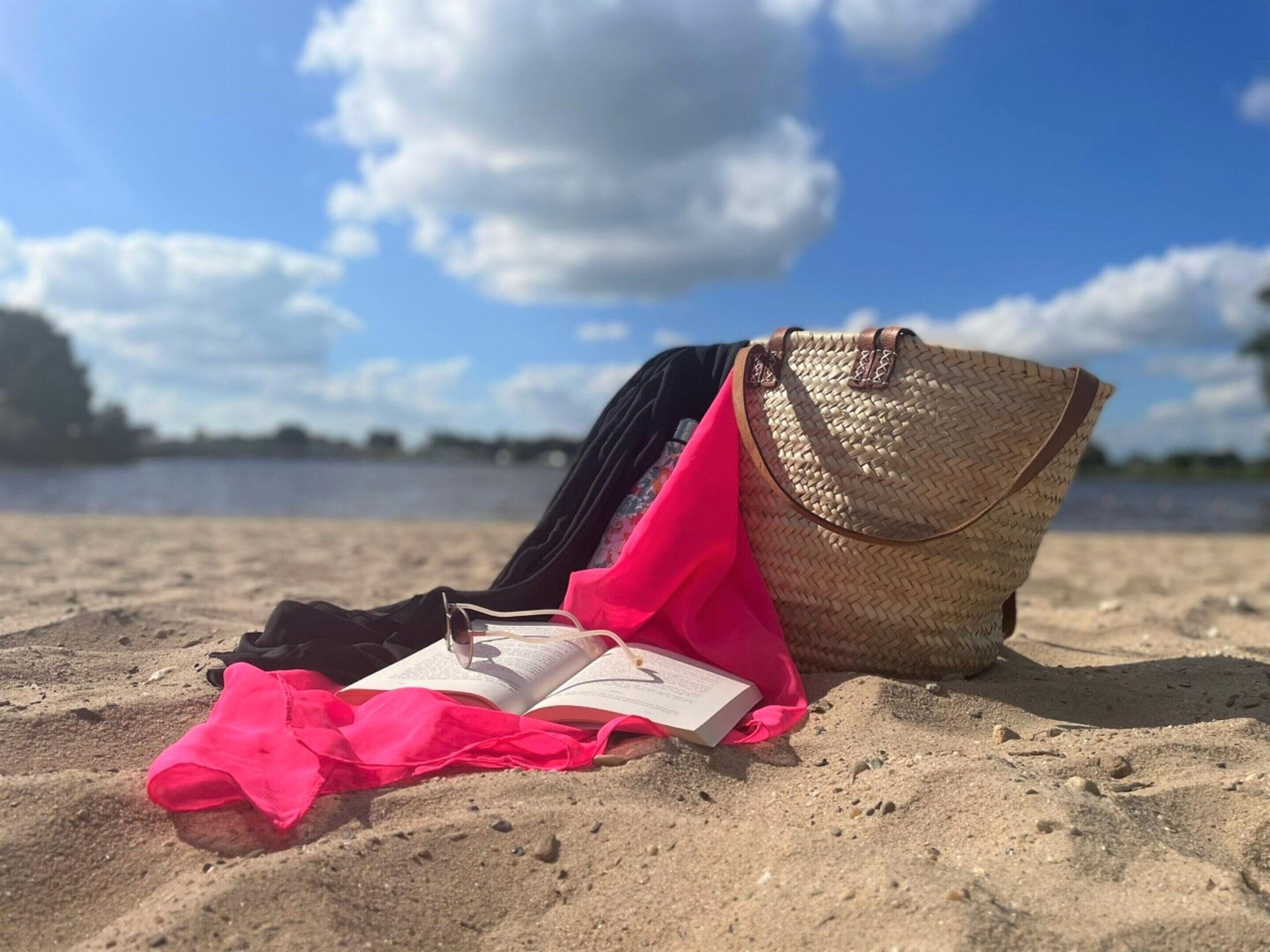 Een strandscène met een rieten tas, een roze doek en een open boek op het zand.