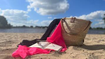 Een strandscène met een rieten tas, een roze doek en een open boek op het zand.