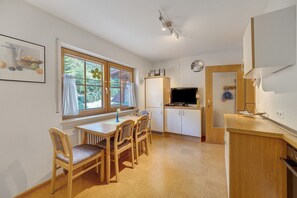 Een kleine keuken met een eethoek, houten keukenkastjes, een ingebouwde televisie en een raam met gordijnen.