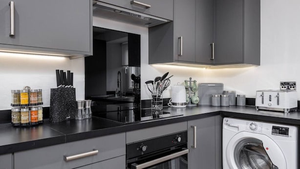 Una cocina moderna con armarios grises, encimera negra, horno empotrado, lavadora y diversos utensilios y electrodomésticos.