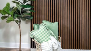 Una habitación con un televisor de pantalla plana grande montado en una pared de madera, una planta en maceta, una silla de mimbre con cojines y una manta tejida.