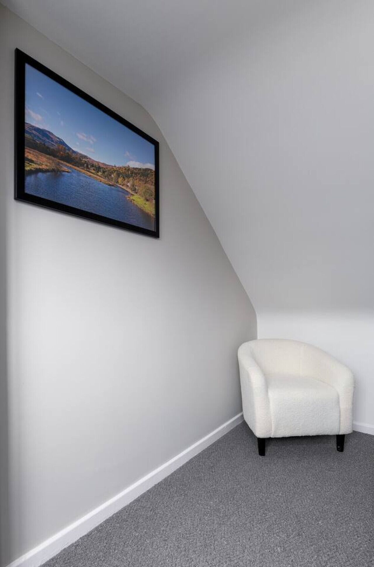 A corner room with a white armchair, a framed landscape painting, and a grey carpet.