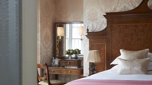 A hotel room with a wooden headboard, a bedside table with a lamp, a mirror, and a dresser with a mirror. - The Goring (London)