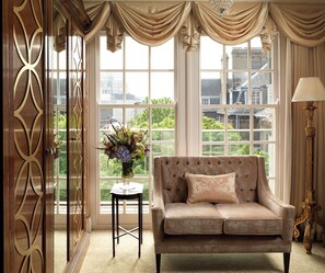 A room with a sofa, a table with a vase, and a view of buildings through large windows. - The Goring (London)