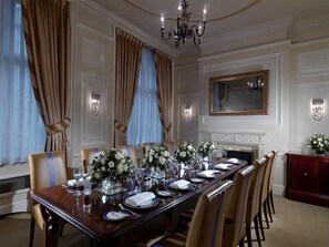 Une salle à manger élégante, avec une longue table dressée pour un repas, entourée de chaises, d’un lustre et d’une cheminée.