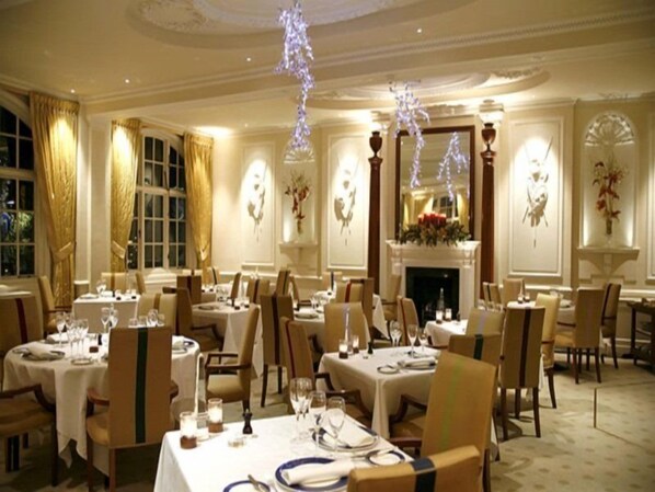 A formal dining room with tables set for multiple guests, elegant chairs, and a fireplace.