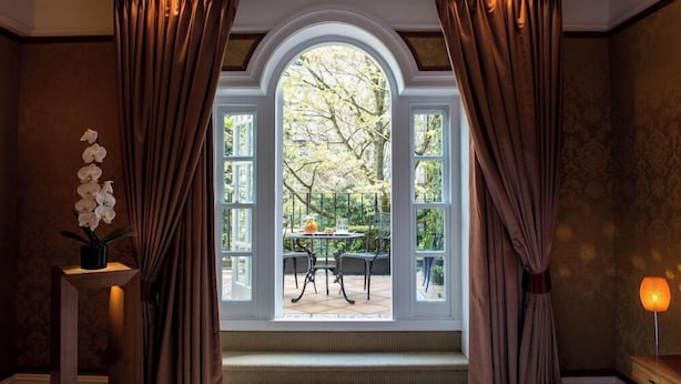 A room with a large arched window, curtains, a potted orchid, a table with chairs, and a lamp. - Goring Hotel (London)