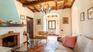 A living room with a fireplace, a glass coffee table, a wicker chair, a sofa with a red pillow, and wooden ceiling beams. - Holiday Home 'Uva' with Guest-Only Pool, Wi-Fi and Air Conditioning (San Gimignano)