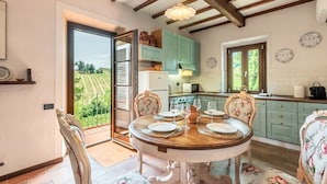 A dining area with a round table set for four, a kitchen with teal cabinets, and a view of a vineyard through a glass door. - Holiday Home 'Uva' with Guest-Only Pool, Wi-Fi and Air Conditioning (San Gimignano)