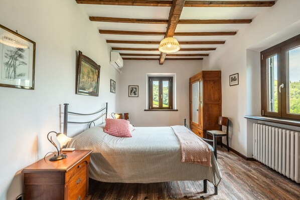 Un dormitorio con cama, mesita de noche, lámpara, ventana y vigas de madera en el techo.