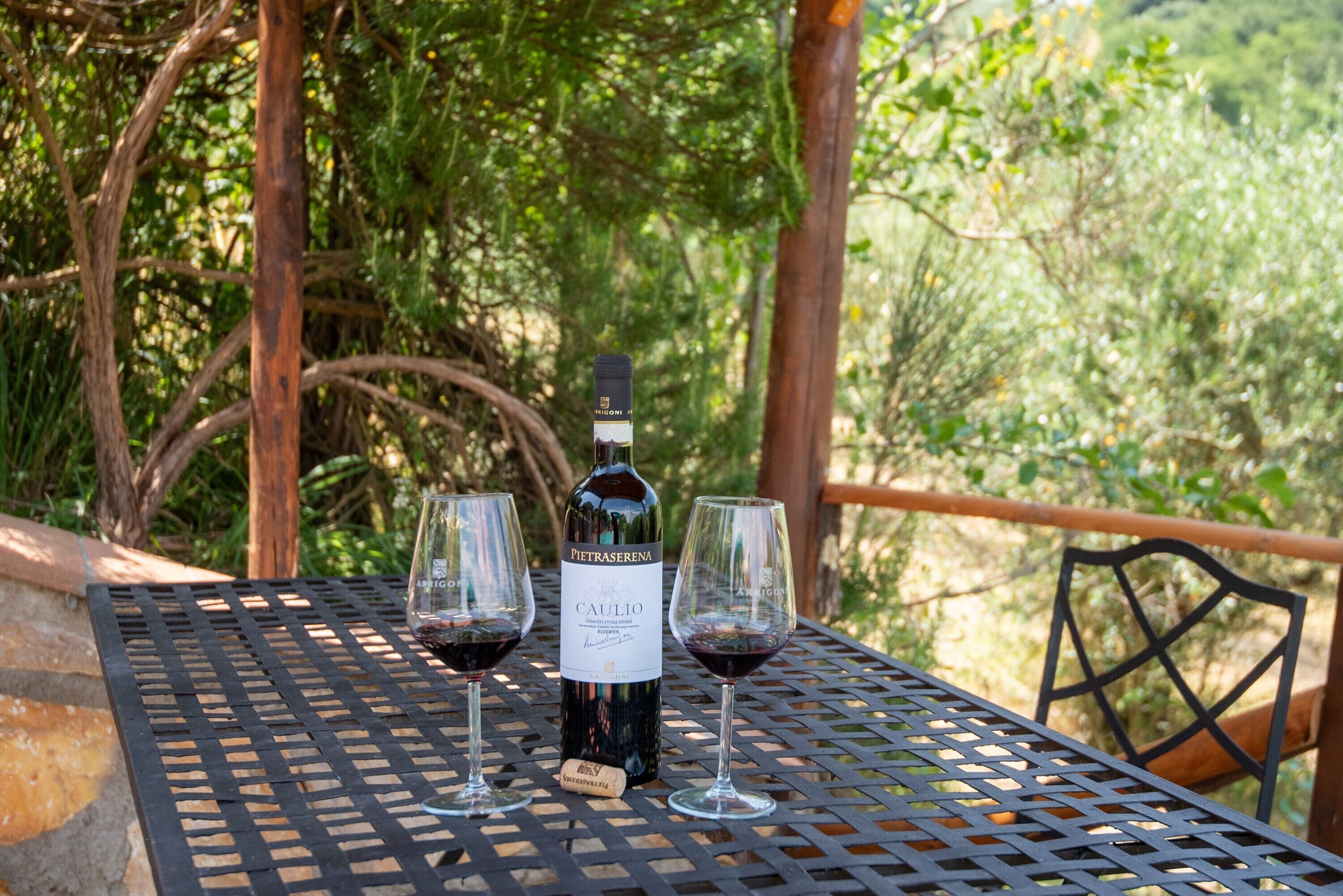 A bottle of Caulio wine and two glasses on a metal table outdoors.