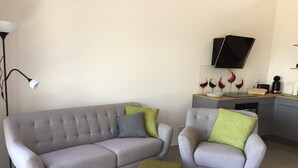 A modern living room with a grey sofa, a matching armchair, and a green ottoman. A flat-screen TV is mounted on the wall above a kitchen counter. - Apartment '2 Spätburgunder' with Balcony and Wi-Fi (Freiburg im Breisgau)
