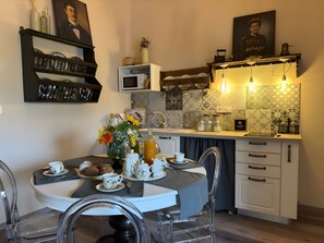 A dining area with a table set for two, a vase with flowers, and a kitchen in the background with a microwave and cabinets.