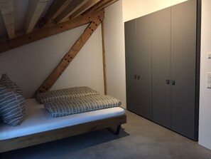 Une chambre à coucher aménagée dans un espace mansardé, avec un plafond en pente, un lit agrémenté d’oreillers à motifs et une grande armoire.