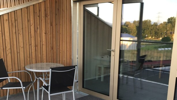 Eine Terrasse mit rundem Tisch und vier Stühlen, einer Schiebetür und Blick auf ein Gebäude und Bäume.